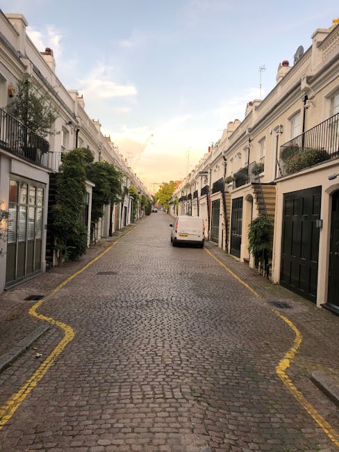 A residential street scene captured during late afternoon or early evening, featuring a row of white Victorian-style terraced houses with decorative facades and front gardens enclosed by white picket 