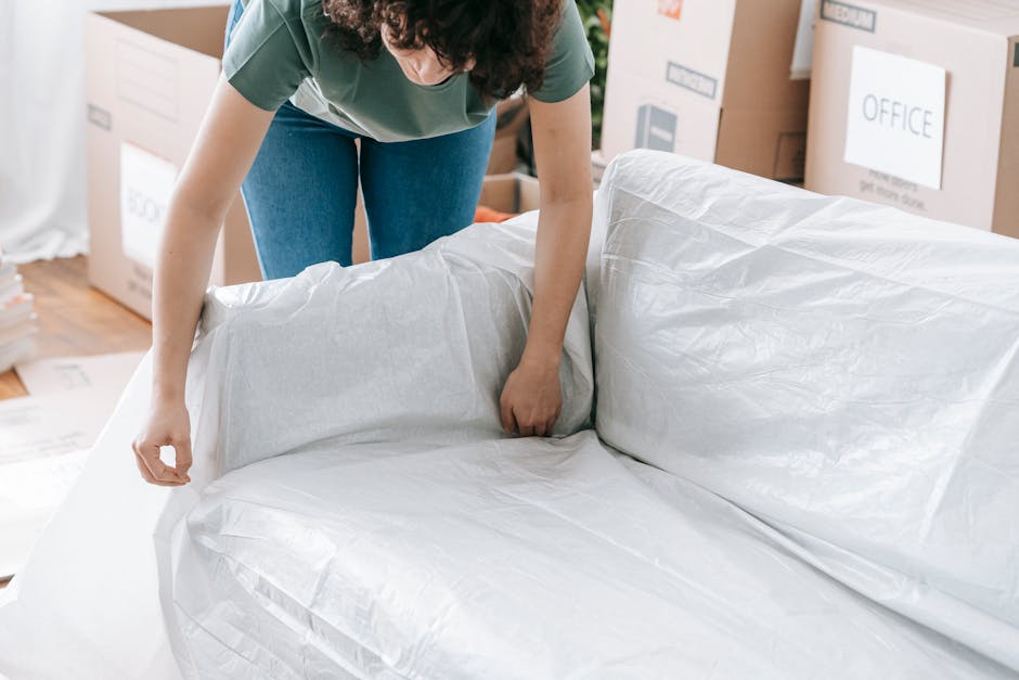 A person wearing a green top and blue jeans is preparing a large piece of furniture, possibly a sofa, by wrapping it in white packing paper or protective plastic sheeting in a well-lit room filled wit